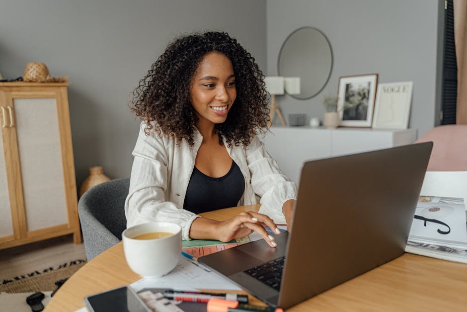 A woman sits at a round table, working on a laptop with a coffee cup nearby in a cozy home office setting.