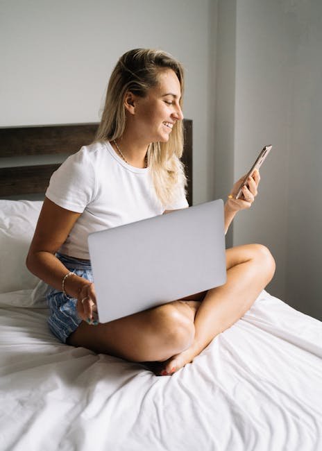 Caucasian woman smiling while using smartphone and laptop at home.